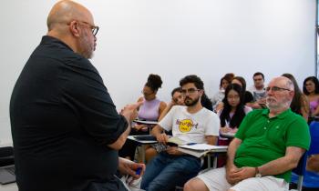 Na fotografia, um professor branco se encontra à esquerda. Ele está de frente para diversos estudantes sentados em carteiras.