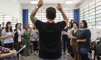 Foto de um grupo de coral em sala de aula sendo regido por um homem de costas para a foto. O regente está de braços erguidos, juntando o indicador e o polegar nas duas mãos. Ele é branco, tem cabelos castanhos curtos, usa uma camiseta preta, um relógio no braço esquerdo, que é parcialmente coberto por tatuagens. À frente do regente há um grupo cantando, tendo fileiras femininas à frente e masculinas ao fundo. Do lado esquerdo da imagem, uma pessoa sentada faz anotações em um papel sobre estante de partitura