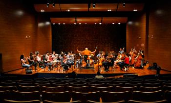 Foto de uma orquestra de câmara em um palco. São cerca de 30 jovens músicos sentados e distribuídos em semicírculo, a maioria com instrumentos de cordas. O regente está sentado em uma cadeira sobre um tablado e tem os braços erguidos, com a batuta na mão direita. Ele está de costas para plateia, que está praticamente vazia. Todos usam roupas coloridas e informais.