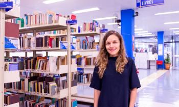 Fotografia de Lilian Viana, mulher branca de cabelos longos e loiros, que veste uma blusa azul escuro e usa um relógio no pulso direito. Ao fundo, há uma biblioteca com prateleiras repletas de livros. Há uma placa no canto superior direito escrito: Livros.