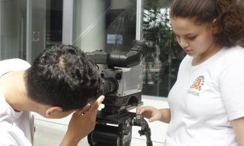 Foto de dois jovens operando uma filmadora. À esquerda da imagem, há uma pessoa branca, de cabelos curtos e pretos, de camiseta branca, de costas e inclinada, mexendo em uma filmadora. A filmadora é preta e cinza, está sobre um tripé preto. À direita da imagem, há uma pessoa branca, de cabelos castanhos amarrados, camiseta branca com o brasão da cidade de Santos, de pé, também mexendo na filmadora. Ao fundo, há janelas transparentes.