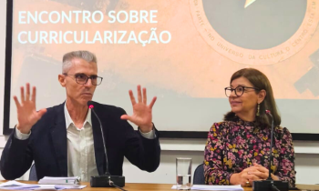 Foto em plano médio. À esquerda, homem branco, de cabelos curtos e grisalhos, usando óculos de grau de haste escura e uma camisa branca quadriculada e um blazer preto. Ele está sentado, ergue as duas mãos para cima, na altura do ombro, enquanto fala ao microfone. Ele é observado por uma mulher que está sentada à direita. Ela é uma mulher branca, de cabelos castanhos, lisos e na altura dos ombros. Ela usa óculos de haste escura e arredondada, brincos dourados, e um vestido preto de manga longa com estampa.