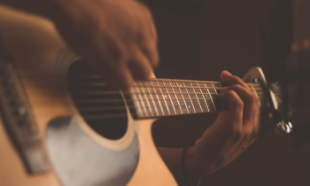 Foto das mãos de uma pessoa tocando um violão de cores claras. Uma das mãos segura o braço do violão, enquanto a outra dedilha as cordas. O fundo é escuro.