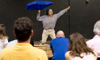 Foto de uma performance em que uma pessoa está com uma máscara dourada de rosto triste. Com a mão direita ela segura um guarda-chuva azul e estende a esquerda na diagonal alta. Ela usa camisa cinza, calça bege e se apresenta diante de uma pequena platéia de costas para a foto. O cenário é preto.