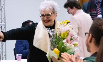 Foto de Maria Immacolata Vassallo de Lopes, uma mulher idosa, de pele e cabelos brancos lisos e curtos, óculos de armação escura e batom vermelho. Ela usa um casaco preto de gola creme e segura um buquê de flores em tons de rosa e amarelo. Ela sorri e acena enquanto algumas pessoas batem palmas no canto inferior direito da imagem. Ao fundo, algumas pessoas estão de pé e sorriem. 