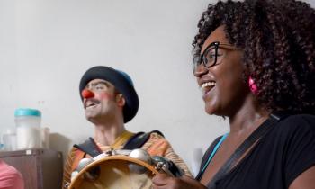 Na foto, um homem branco à esquerda e uma mulher negra à direita tocam instrumentos e cantam sorridentes. O homem usa chapéu azul, nariz de palhaço vermelho e blusa laranja listrada. A mulher, que está em primeiro plano, tem cabelos cacheados castanhos até a altura dos ombros, usa óculos de armação preta e camiseta preta. Ela segura um pandeiro que encobre o instrumento dele e ambos estão voltados para o lado esquerdo da imagem. Ao fundo, uma parede branca e um armário, sobre o qual há alguns potes plástico