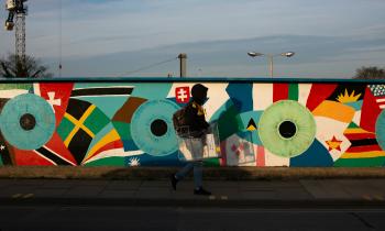 Foto de uma pessoa caminhando em uma calçada, tendo ao fundo um muro grafitado. O grafite é composto por círculos grandes azuis ou verdes que têm em seu centro círculos pretos. Em volta deles, há formas coloridas como retalhos de bandeiras de países. A pessoa  usa um moletom preto com capuz cobrindo o rosto, calça jeans, tênis, e carrega uma mochila e um objeto de metal. Atrás do muro, há o topo de duas árvores e um poste, sob um céu azul.