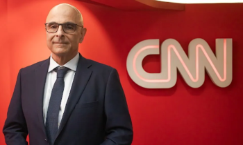 Foto de um homem branco sorrindo. Ele veste terno e gravata azuis, óculos de armação quadrada e preta. Ao fundo, uma parede vermelha e um letreiro com o texto “CNN”.