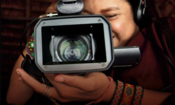 Foto de uma pessoa indígena segurando uma câmera filmadora preta em frente ao rosto. A lente da câmera apoiada em uma das mãos da pessoa aparece em primeiro plano. Ela tem cabelos escuros, sorri, usa um headfonepreto e pulseiras de miçangas coloridas nos punhos. O fundo da imagem é escuro.