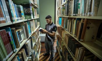 Foto de um rapaz folheando um livro em uma biblioteca.  Ele usa óculos, tem cabelos curtos e pretos, veste roupa esportiva e está entre dois corredores de estantes cheias de livros.