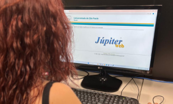 Fotografia de uma mulher olhando a tela de um computador. A mulher está de costas para a câmera, tem pele branca e cabelo vermelho e observa a tela do computador onde é há uma logo escrita “Júpiter web”. 