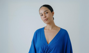 Na foto, uma mulher negra com cabelos pretos presos em um coque baixo. Ela veste blusa azul com decote V e usa brincos. O fundo é cinza.