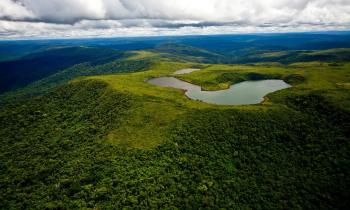 Foto panorâmica da Floresta Nacional de Carajás vista de cima. Em primeiro plano, há um grande morro coberto por vegetação e, no topo dele, um lago. Em segundo plano, há um mar de morros também cobertos por vegetação, sendo que quanto mais distante estão, mais escuros e azulados. Acima da linha do horizonte, o céu nublado. 