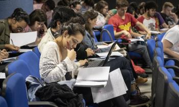 Foto de um grupo de estudantes em um auditório. Em primeiro plano, destaca-se uma pessoa de pele clara e cabelos presos que olha para um caderno com o queixo apoiado na mão direita, que também segura uma caneta. As cadeiras do auditório são azuis e a maioria está ocupada com pessoas que olham ou manuseiam cadernos e folhas de papel. 