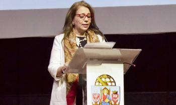 Foto de uma mulher branca, que discursa, de pé, atrás de um púlpito. Ela tem cabelos longos e loiros e usa óculos com armação vermelha, veste um blazer branco e tem um lenço estampado em volta do pescoço. Ela olha ligeiramente para baixo, para ler um papel a sua frente. Atrás dela, um fundo preto e cinza. 