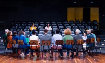 Fotografia de um grupo de doze pessoas sentadas em cadeiras de madeira em semicírculo no palco de um teatro. De frente para elas está a plateia com apenas duas pessoas sentadas. As pessoas nas cadeiras estão de costas para a câmera e têm diversas aparências e cores de vestimenta. O piso é de madeira, as paredes do teatro são escuras e, ao fundo, há duas portas de incêndio amarelas fechadas.