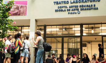 Foto de grupo de aproximadamente 20 estudantes em frente ao Teatro Laboratório da ECA. Do lado esquerdo da foto, há um grupo em pé e conversando. Do lado direito, há pessoas sentadas no chão. A fachada do prédio possui um letreiro azul onde está escrito Teatro Laboratório, Depto. Artes Cênicas e Escola de Arte Dramática, Prédio 8. Possui paredes bege e portas de vidro com esquadrias pretas.