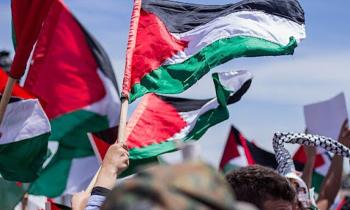 Foto de uma manifestação na Palestina. No centro, é possível ver o braço de uma pessoa segurando um mastro com uma bandeira da Palestina, que tem uma linha horizontal preta, uma branca e uma verde e um triângulo vermelho à esquerda. À esquerda e à direita há mais pessoas segurando a mesma bandeira. Ao fundo, há um céu azul.