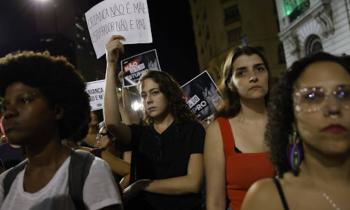 Foto de uma manifestação contra a PL 1904/2024. No centro, há uma mulher branca, de cabelos ondulados, castanhos e longos, e veste camiseta preta. Ela segura um cartaz com os dizeres “Criança não é mãe, estuprador não é pai”. À esquerda, à direita e atrás há mulheres protestando e segurando cartazes. Ao fundo, há prédios e está de noite.