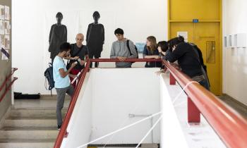 Foto de um grupo de sete pessoas que conversa apoiado em um corrimão vermelho em formato de U. No centro, se destaca o vão criado pela escada, que aparece à esquerda. À direita, um corredor e uma porta amarela. Ao fundo, uma parede branca com reproduções em grande formato de fotos em preto e branco de duas pessoas. 