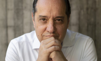Foto de um homem branco, calvo, de cabelos escuros e curtos. Ele veste camisa social branca e relógio de pulso prateado. Sorri ligeiramente, com as mãos unidas sob o queixo e os cotovelos apoiados sobre uma mesa. O fundo é cinza e desfocado.