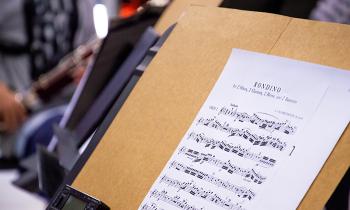 Foto da partitura da música Rondido. A página impressa está apoiada num suporte de madeira com um metrônomo apoiado no canto inferior esquerdo. Ao fundo, desfocada, do lado esquerdo, há uma pessoa tocando um instrumento de sopro.