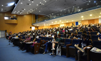 Foto de diversas pessoas sentadas na plateia de um grande auditório. O chão tem carpete azul e as paredes são de madeira clara. No teto branco há diversas lâmpadas amarelas e brancas. Alguns microfones estão posicionados em pedestais diante da primeira fileira de assentos.