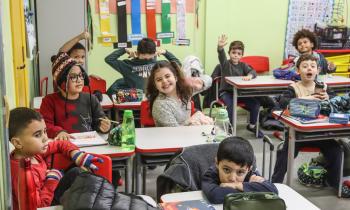 Foto de uma sala de aula. Há nove crianças sentadas em suas carteiras brancas e cadeiras vermelhas, enfileiradas em duplas. Algumas sorriem para a foto e outras olham em outras direções. Elas vestem roupas de frio. As paredes são verdes, com cartazes e materiais educativos e coloridos pendurados.