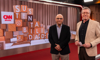 Foto de dois homens brancos à frente de uma tela digital na qual aparece o logo CNN Brasil e letras pintadas em blocos de madeira empilhados formando a palavra Sustentabilidade. O homem à esquerda é careca, usa óculos de armação quadrada e preta, camisa azul, blazer preto e calça marrom. O homem à direita usa camisa preta, blazer bege, calça preta, óculos de armação redonda e marrom e tem cabelos curtos e grisalhos. 
