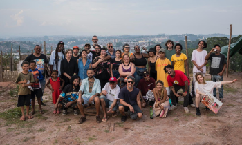 Na foto, um grupo de cerca de 30 pessoas olha para a câmera e sorri em um espaço aberto com chão de terra. Elas têm diferentes idades, expressões de gênero, identificação racial e vestimentas. Ao fundo, a vista panorâmica de uma cidade e o céu azul. 