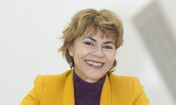 Foto de uma mulher branca, de cabelos loiros, curtos e lisos. Ela veste uma blusa roxa e um paletó amarelo. Ela está com as mãos uma sobre a outra e apoiadas sobre uma mesa. O fundo é branco.