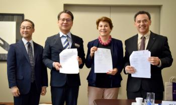 Foto de quatro pessoas, de pé, posando e sorrindo. Da esquerda para direita estão: dois homens amarelos, vestindo terno e gravata, uma mulher branca, de cabelos curtos, lisos e castanhos, vestindo um blazer azul marinho e um homem pardo, vestindo terno e gravata. Dois homens e a mulher seguram com as duas mãos uma folha de papel cada com as assinaturas do convênio. Ao fundo, uma parede bege. 