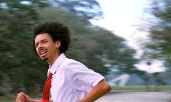 Foto de homem jovem negro correndo. Seus cabelos são escuros, de tamanho médio e cacheados, ele tem cavanhaque e bigode,  veste camisa social branca com gravata vermelha e usa brinco na orelha. 