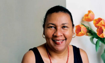 Foto de mulher negra de olhos castanhos e cabelos escuros e presos sorrindo. Ela veste regata preta, usa colar de contas vermelhas e brincos de argolas douradas. No fundo, uma parede cinza e branca e flores cor de laranja à direita.