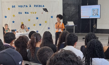 Foto de uma mulher negra falando ao microfone para um grupo de pessoas sentado à sua frente e de costas para a câmara. Ela está de pé, tem cabelos crespos que estão presos, usa uma camiseta laranja e uma calça preta. Atrás dela, uma mesa com duas mulheres e um painel branco onde está escrito “De volta à Escola”, “Eu na USP”, decorado com várias estrelas, em amarelo e azul. Atrás dela também há uma televisão que mostra um código QR. 