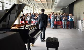 Foto de um pianista de pé diante de uma pequena plateia. O piano de cauda está em primeiro plano, à esquerda, e o pianista, usando terno, está ao centro e de costas para a câmera. Ao fundo, diversas pessoas, na maioria brancas, está de pé e batem palmas. As cadeiras do local são vermelhas e se destacam sobre o piso de concreto claro. 
