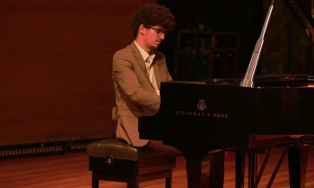 Foto de um homem negro de pele clara, sentado em um banco e tocando piano de cauda. Ele tem cabelos crespos, castanhos e curtos, veste um terno marrom com camisa branca e usa óculos de armação preto. O fundo é escuro.
