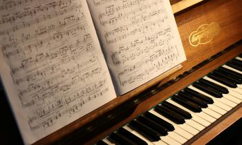 Foto de um piano com um livro de partituras aberto apoiado em seu suporte. O piano é de madeira, e parte do teclado com teclas pretas e brancas está visível. A partitura contém várias notas e anotações manuscritas. A luz ilumina a partitura e parte do piano.