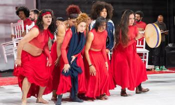 Foto de uma apresentação teatral em um palco. Um grupo de pessoas vestidas com roupas vermelhas está no centro da cena, olhando para a frente. Algumas estão em postura curvada e uma delas toca um tambor de mão. Ao fundo, outras pessoas, também vestidas de vermelho, seguram cadeiras brancas. O piso é claro e as paredes são escuras. 