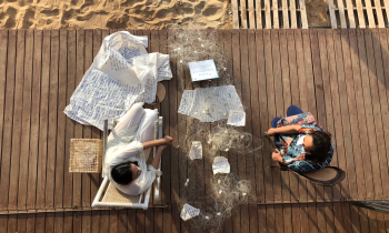 Foto de cima de duas mulheres sentadas em cadeiras sobre um piso de madeira. Ambas são brancas, de cabelos castanhos, mas uma delas aparenta ser mais jovem que a outra. Entre elas, há uma rede de pesca com papéis escritos dispostos sobre ela. Ao lado da mulher sentada à esquerda, um grande tecido claro coberto por escritos em azul.