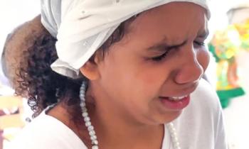Foto de menina negra de olhos fechados e com expressão emocionada. Ela veste um turbante branco, tem cabelos castanhos escuros cacheados na altura do ombro e usa um fio de contas brancas e camiseta branca. O fundo é claro. 