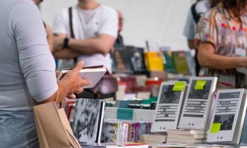 Foto de pessoa segurando livro em frente a uma bancada cheia de livros. Ao fundo estão livros e pessoas em desfoque.