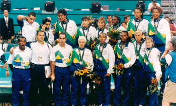  Foto da seleção brasileira feminina de basquete posando com medalhas de prata. Do lado esquerdo, o técnico e outros homens da comissão técnica. Os homens vestem calças pretas e azuis e camiseta branca com detalhes em verde, amarelo e azul. As mulheres vestem calça azul e agasalhos brancos com detalhes em verde, amarelo e azul. Uma pessoa à frente segura uma câmera de vídeo e veste bermuda bege e camiseta azul. Ao fundo está uma arquibancada e algumas pessoas.