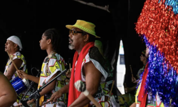 Foto de um grupo de pessoas negras de perfil, sendo que três estão em foco e seguram baquetas e tambores. Elas usam roupas coloridas de branco, amarelo, preto, vermelho e verde. Uma pessoa usa boina branca e outra um chapéu amarelo. O fundo é escuro e, no canto direito da imagem, há franjas vermelhas e azuis de papel celofane. 