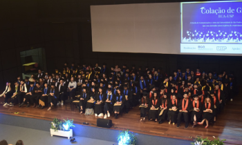 Foto de cerca de 150 estudantes, vestindo becas e sentados lado a lado em cadeiras posicionadas no palco de um auditório. Ao fundo, uma tela de projeção, que exibe arte em que se lê, parcialmente, “Colação de grau da ECA-USP”. À frente do palco, há três arranjos de flores.