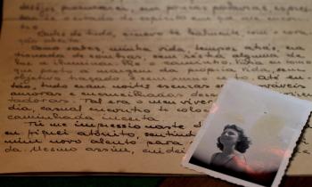  Em uma superfície, uma carta com escritos em letra cursiva e tinta preta e uma fotografia em preto e branco de uma mulher sobre a carta, no canto direito da imagem. A mulher tem cabelos curtos ondulados e sorri olhando na direção de algo que não aparece na foto. A carta é amarelada e a fotografia está com manchas amareladas também. 