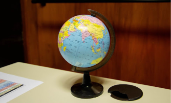 Foto de um pequeno globo terrestre que mostra principalmente o continente asiático, com países nas cores amarelo, rosa, laranja e verde, e o oceano Índico em azul. O globo está sobre uma mesa clara com detalhes em preto. Sobre a mesa há também um papel colorido. O fundo, desfocado, é uma porta de madeira. 