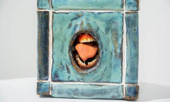 Foto de escultura em cerâmica de um cubo com uma boca aberta no centro. O cubo é azul e sua estrutura lembra pisos de cerâmica com rejunte branco. A boca é um buraco no centro do cubo e exibe língua, dentes e gengivas. 