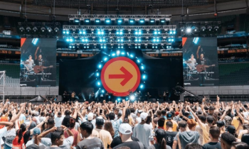 Foto de show musical com centenas de pessoas. A maioria do público é jovem e veste roupas claras. No palco, a banda e, ao fundo, um cenário com refletores e a imagem de uma seta vermelha em um círculo laranja, apontando para a direita. Acima, refletores com a cor azul. O palco é ladeado por telões que exibem imagens da banda.