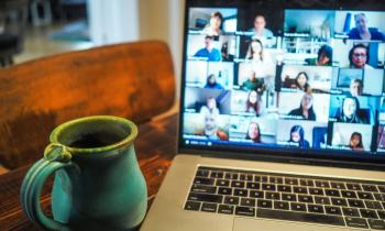 Foto de notebook sobre uma mesa. Na tela, há diversas imagens de pessoas em uma reunião online, ao lado do computador há uma caneca verde. 